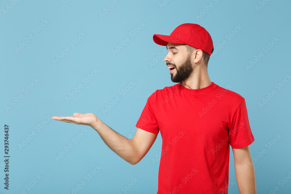 Delivery man in red uniform workwear isolated on blue wall background, studio portrait. Professional male employee in cap t-shirt print working as courier dealer. Service concept. Mock up copy space. Delivery man in red uniform workwear isolated on blue wall background, studio portrait. Professional male employee in cap t-shirt print working as courier dealer. Service concept. Mock up copy space.