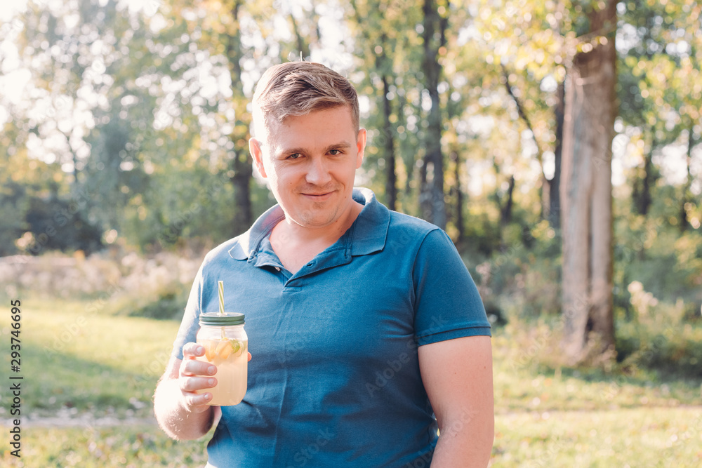 Man in casual wear posing over green city park on sunny warm day, holding cup of lemonade and looking to camera with soft smile. Vitamins and regular life concept Man in casual wear posing over green city park on sunny warm day, holding cup of lemonade and looking to camera with soft smile. Vitamins and regular life concept