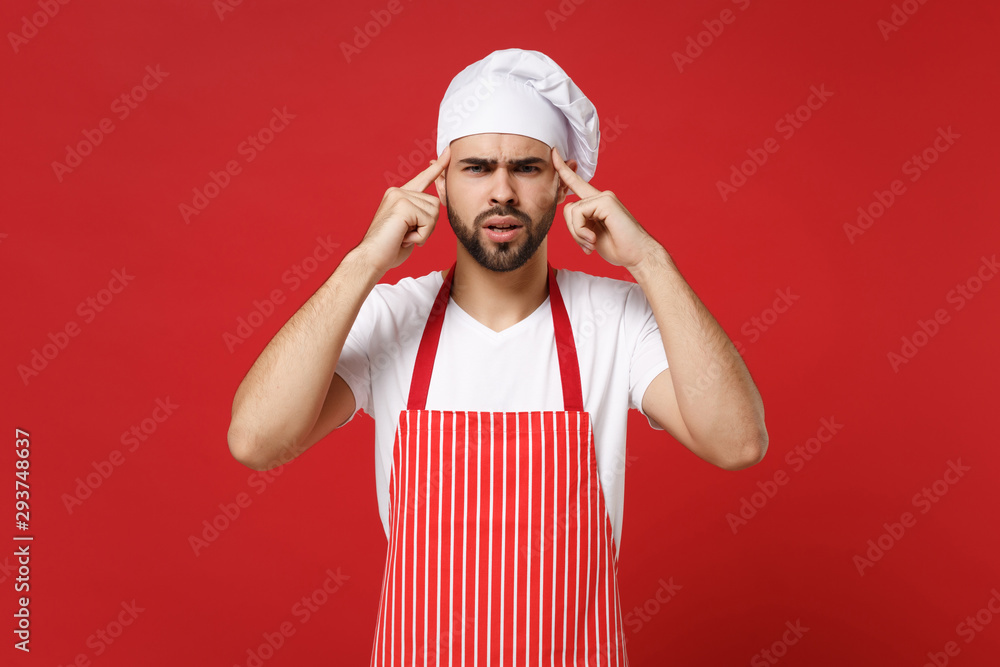 Preoccupied young bearded male chef cook or baker man in striped apron toque chefs hat posing isolated on red wall background. Cooking food concept. Mock up copy space. Pointing index fingers on head. Preoccupied young bearded male chef cook or baker man in striped apron toque chefs hat posing isolated on red wall background. Cooking food concept. Mock up copy space. Pointing index fingers on head.