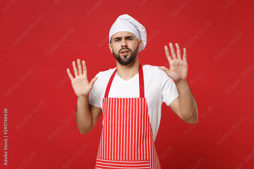 Shocked scared bearded male chef cook or baker man in striped apron t-shirt toque chefs hat posing isolated on red background. Cooking food concept. Mock up copy space. Rising hands, showing palms. Shocked scared bearded male chef cook or baker man in striped apron t-shirt toque chefs hat posing isolated on red background. Cooking food concept. Mock up copy space. Rising hands, showing palms.