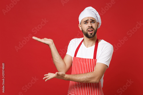 Perplexed young bearded male chef cook or baker man in striped apron white t-shirt toque chefs hat posing isolated on red background. Cooking food concept. Mock up copy space. Pointing hands aside.