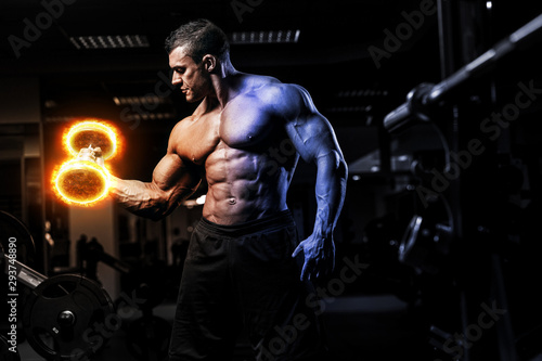 Muscular athletic bodybuilder fitness model training arms with dumbbells on fire in gym. Concept sport photo of exercises in gym