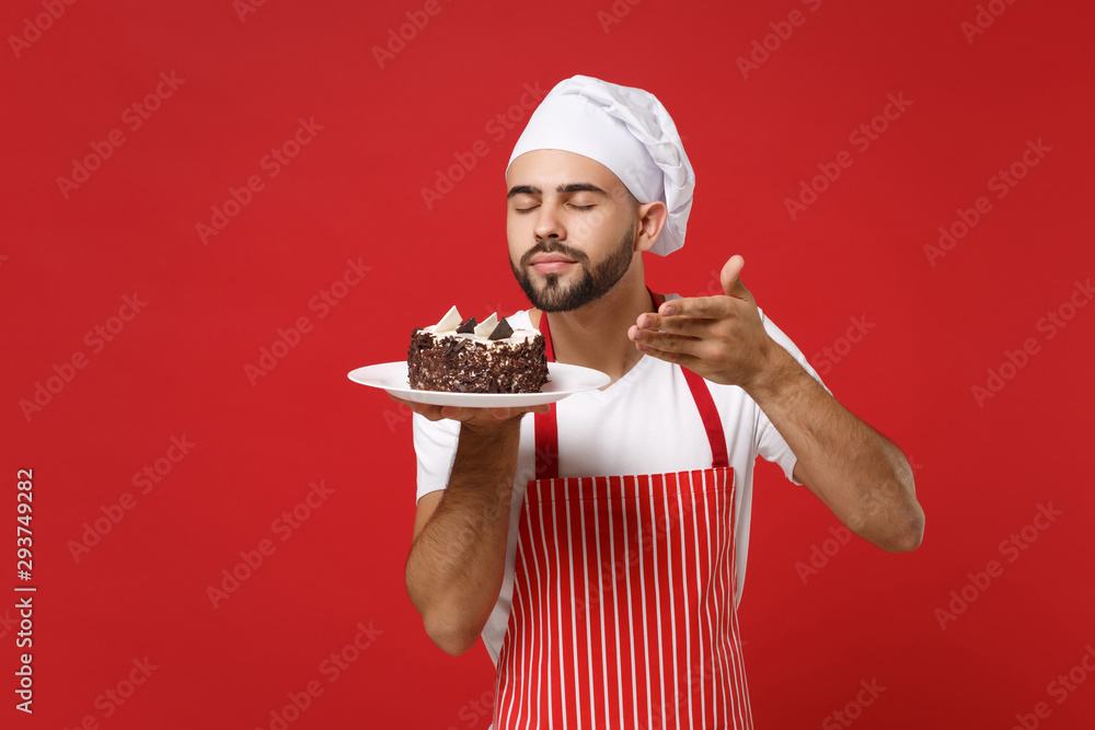 Male chef cook or baker man in striped apron toque chefs hat posing isolated on red background. Cooking food concept. Mock up copy space. Hold plate with cake, raised hand to face feeling food smell.