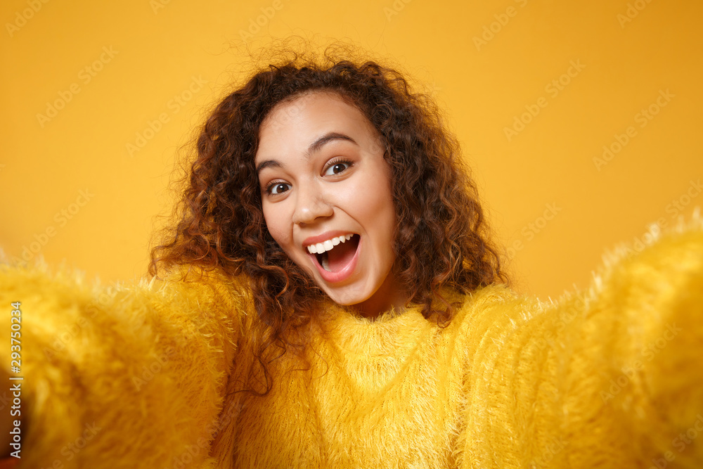 Close up of excited young african american girl in fur sweater posing isolated on yellow orange background in studio. People lifestyle concept. Mock up copy space. Doing selfie shot on mobile phone. Close up of excited young african american girl in fur sweater posing isolated on yellow orange background in studio. People lifestyle concept. Mock up copy space. Doing selfie shot on mobile phone.