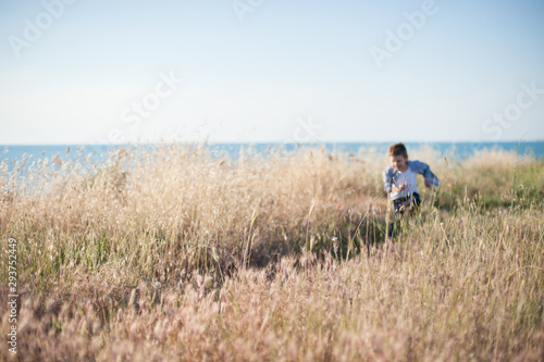 funny happy healthy small caucasian kid running away among dry grass with blue sky and sea horizon on background during outdoor leisure activity