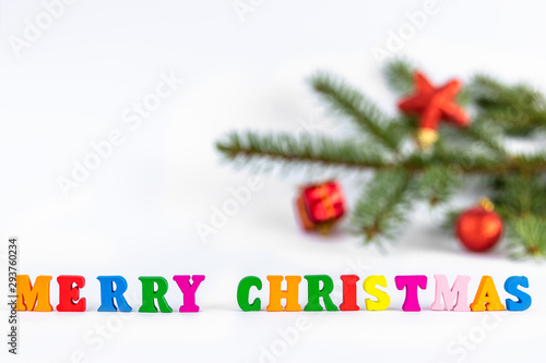 Creative layout of words merry christmas made of wooden multi-colored letters on white background with blurred fir branch and red toys. New Year concept with copy space for note and text