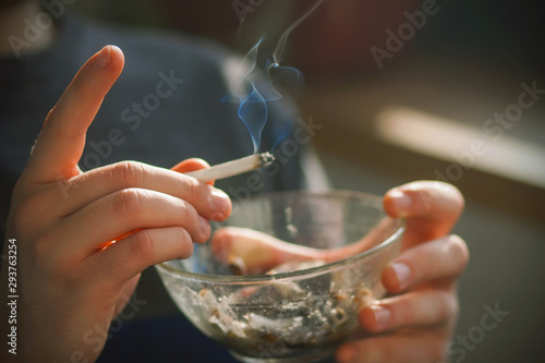 The human holds in his hands, illuminated by bright sunlight, glass dirty ashtray for cigarette butts and shakes into it the ash from the cigarette, which emits blue harmful smoke.