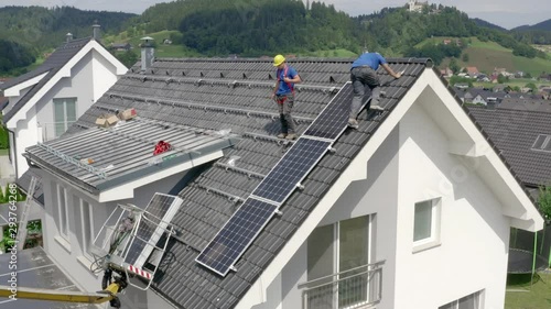 Slow orbital panning drone shot of two workers in blue shirts and yellow helmets mounting solar panels on the roof of twhite house in residential area photo