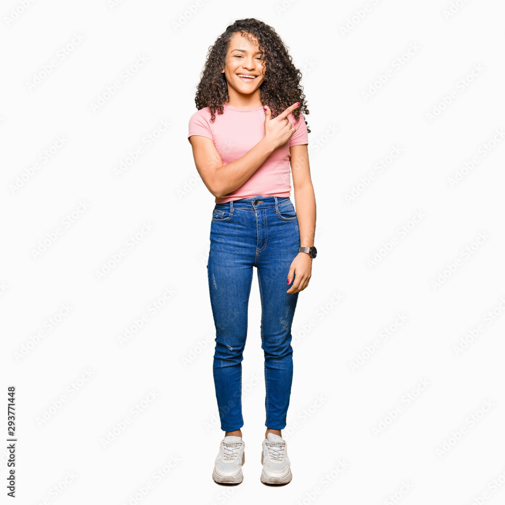 Young beautiful woman with curly hair wearing pink t-shirt cheerful with a smile of face pointing with hand and finger up to the side with happy and natural expression on face Young beautiful woman with curly hair wearing pink t-shirt cheerful with a smile of face pointing with hand and finger up to the side with happy and natural expression on face