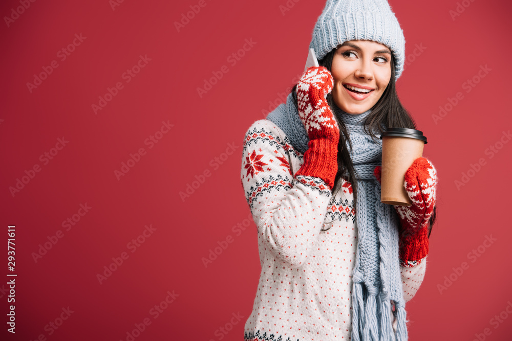 smiling woman in winter clothes talking on smartphone and holding coffee to go isolated on red