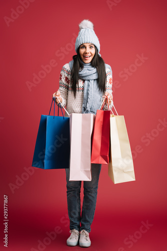smiling woman in winter sweater, scarf and hat holding shopping bags, isolated on red