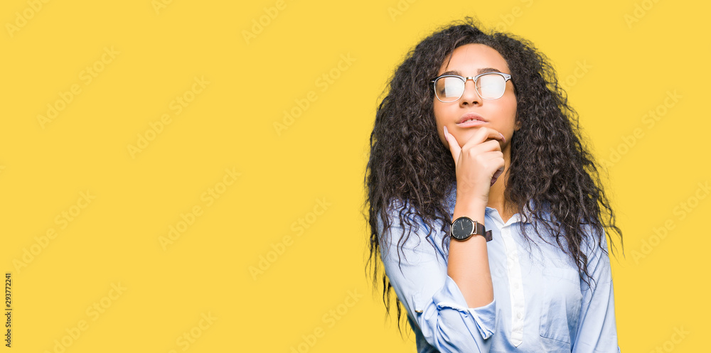 Young beautiful business girl with curly hair wearing glasses looking confident at the camera with smile with crossed arms and hand raised on chin. Thinking positive. Young beautiful business girl with curly hair wearing glasses looking confident at the camera with smile with crossed arms and hand raised on chin. Thinking positive.