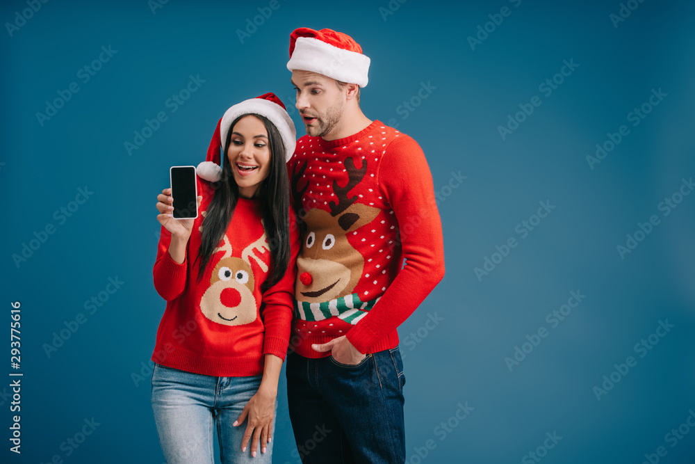 happy couple in santa hats and christmas sweaters showing smartphone with blank screen isolated on blue happy couple in santa hats and christmas sweaters showing smartphone with blank screen isolated on blue