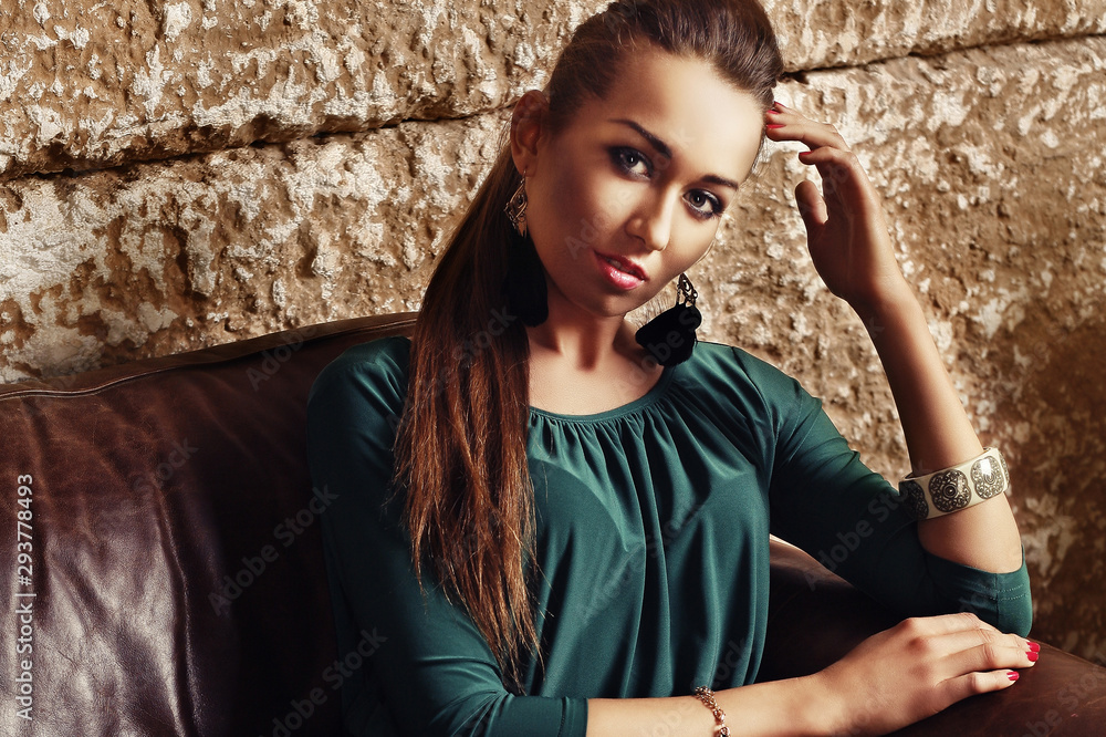 Gorgeous stunning young brunette woman in emerald dress sitting on leather sofa and looking at camera. Fashion model portrait. Gorgeous stunning young brunette woman in emerald dress sitting on leather sofa and looking at camera. Fashion model portrait.