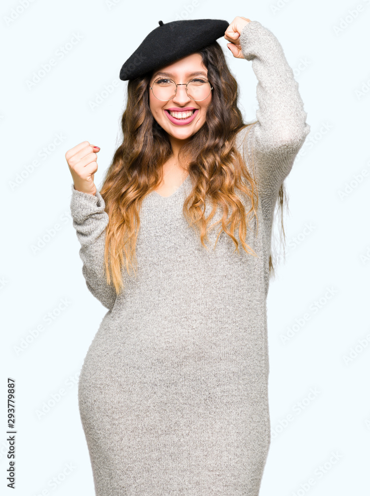 Beautiful young woman wearing glasses and fashion beret very happy and excited doing winner gesture with arms raised, smiling and screaming for success. Celebration concept. Beautiful young woman wearing glasses and fashion beret very happy and excited doing winner gesture with arms raised, smiling and screaming for success. Celebration concept.