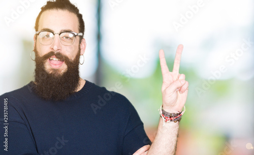 Young hipster man with long hair and beard wearing glasses smiling with happy face winking at the camera doing victory sign. Number two.