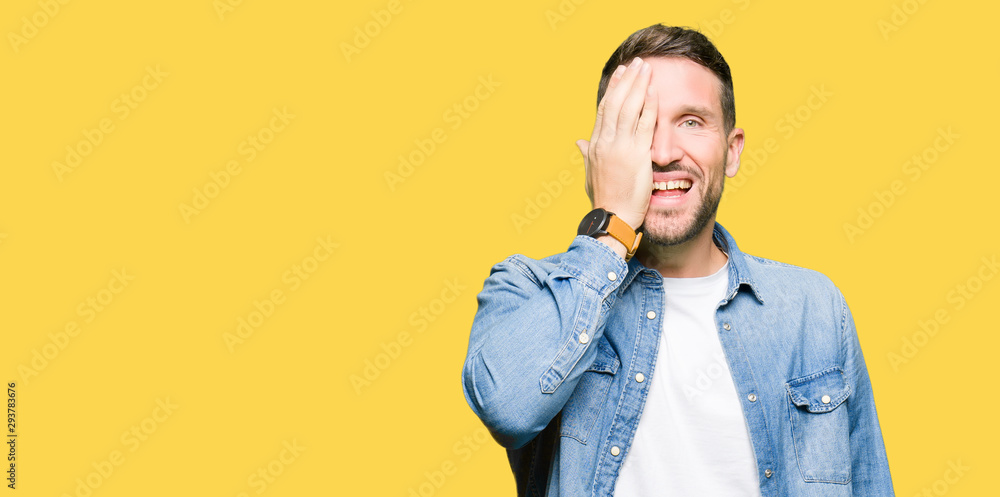 Handsome man with blue eyes and beard wearing denim jacket covering one eye with hand with confident smile on face and surprise emotion. Handsome man with blue eyes and beard wearing denim jacket covering one eye with hand with confident smile on face and surprise emotion.