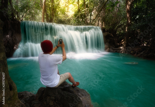 A young asia tourism man sitting in front of a beautiful waterfall in the forest. Happy feeling Full, Travel life, make your asia journey. Taking waterfall photo using smart phone camera