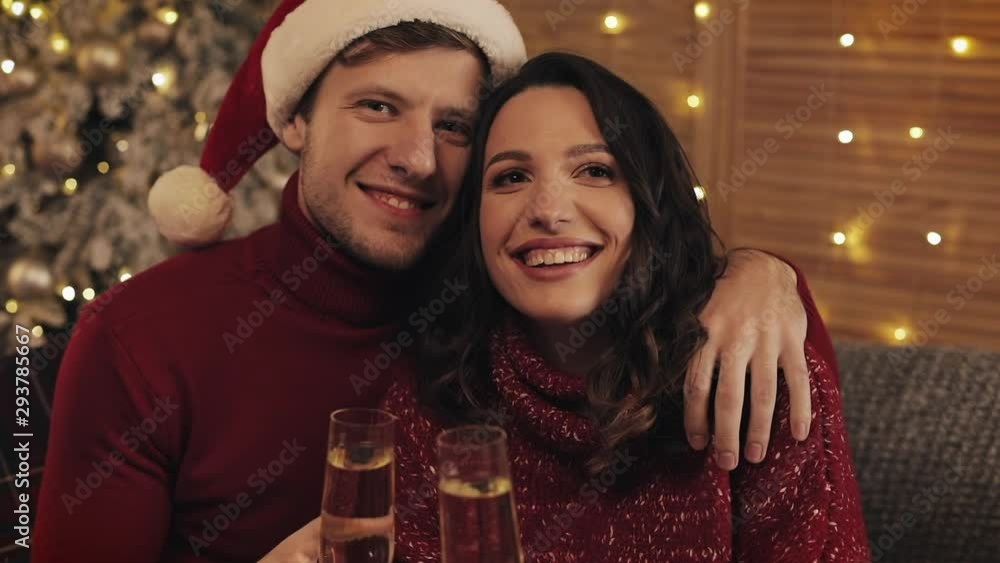 Portrait of Lovely Young Couple Sitting on the Sofa near Christmas Tree at Cosy Home Background Smiling and Raising Glasses of Champagne to Camera Concept of Holidays and New Year. Portrait of Lovely Young Couple Sitting on the Sofa near Christmas Tree at Cosy Home Background Smiling and Raising Glasses of Champagne to Camera Concept of Holidays and New Year.