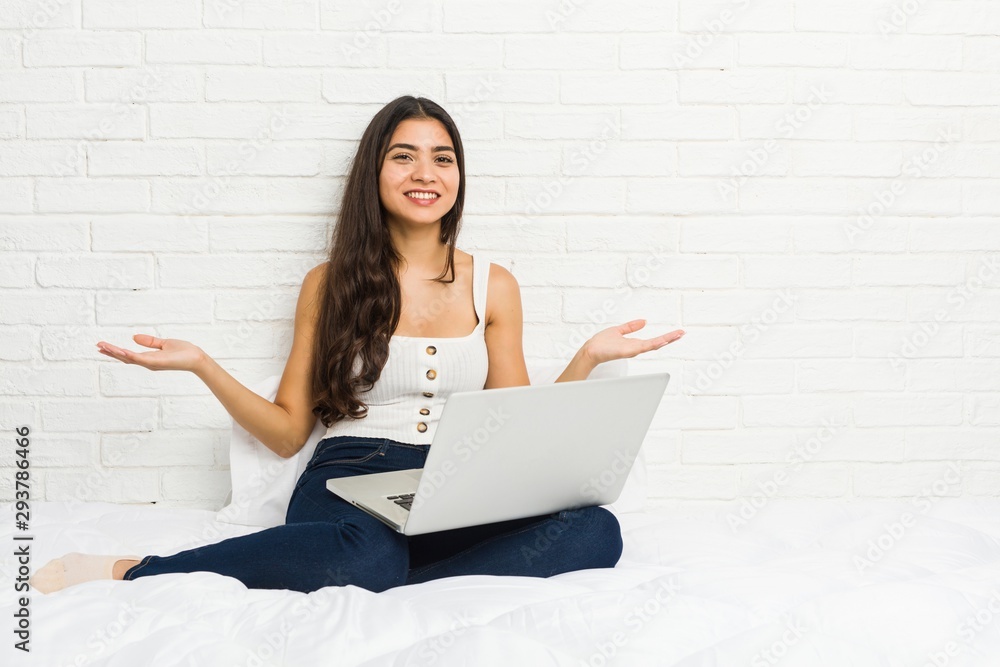 Young arab woman working with her laptop on the bed showing a welcome expression. Young arab woman working with her laptop on the bed showing a welcome expression.
