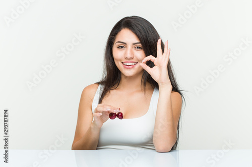 Young hispanic woman holding cherries cheerful and confident showing ok gesture.
