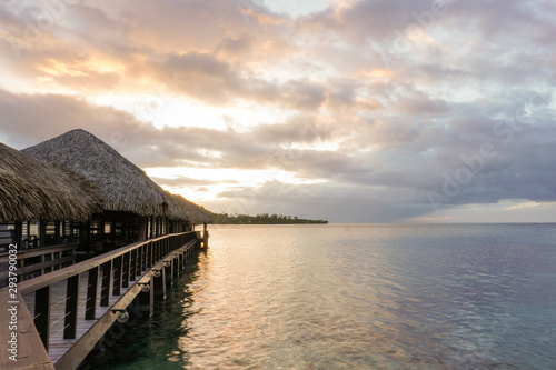 A wooden walkway with thatched roof bungalows leads out into the lagoon on the island of Moorea in French Polynesia at sunset  ocpy space