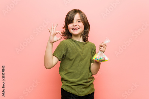 Little boy caucasian holding candies cheerful and confident showing ok gesture.