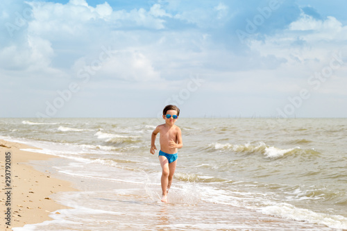 A boy in sunglasses runs along the sea coast on golden sand. Rest of children on summer vacation. Holiday background with copy space. Front view.
