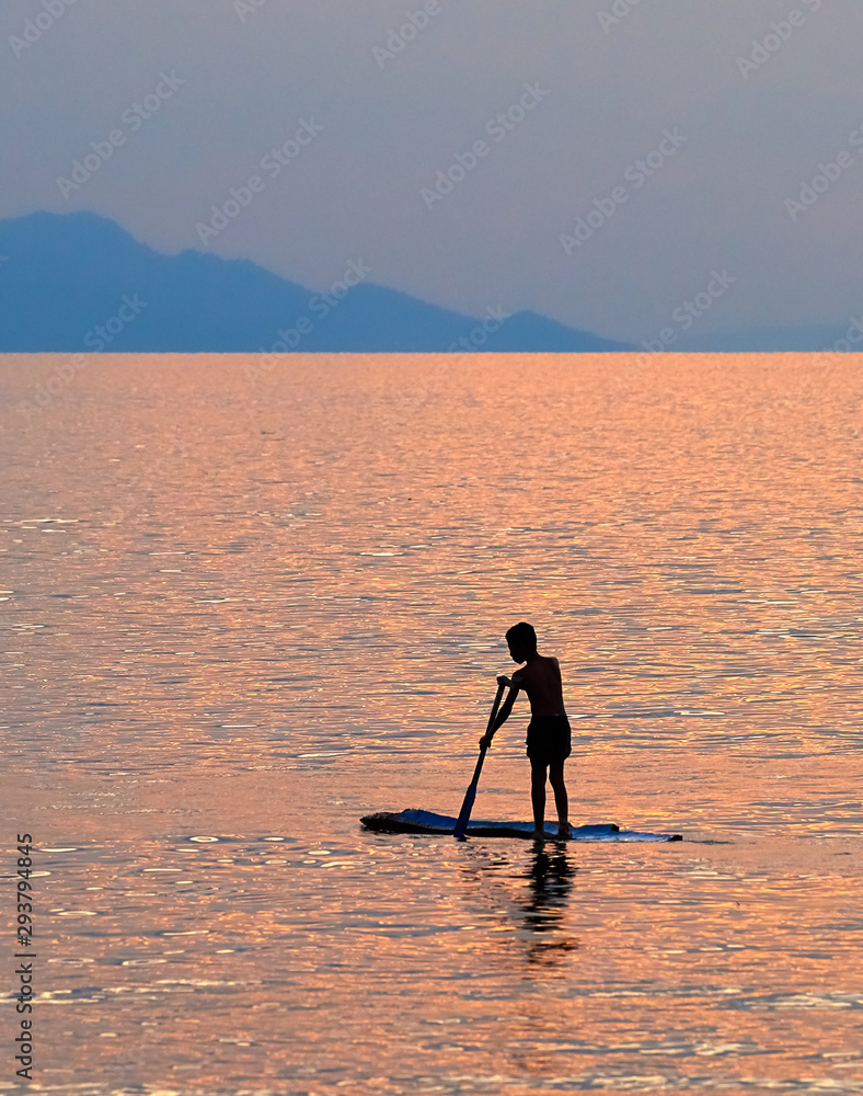 A boy standing on a raft floats on the pink sea from sunset