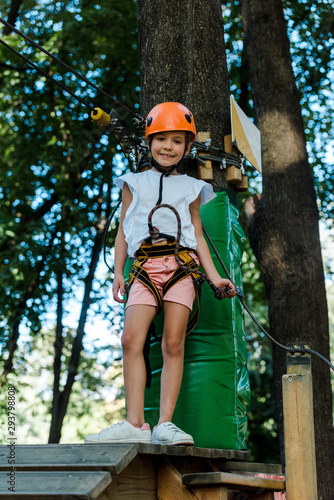 cheerful kid in helmet with height equipment in adventure park near trees