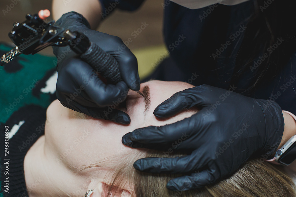 master in black gloves does permanent eyebrow makeup to a young beautiful blonde girl 1