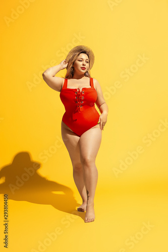 Young caucasian plus size female model's preparing for beach resort on yellow background. Woman in red swimsuit and hat posing confident. Concept of summertime, party, body positive, equality. © master1305