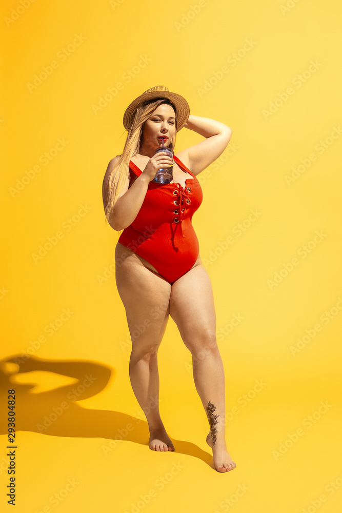 Young caucasian plus size female model's preparing for beach resort on yellow background. Woman in red swimsuit and hat drinking cocktail. Concept of summertime, party, body positive, equality. - obrazy, fototapety, plakaty 