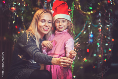 Happy family woman mother and little girl relax playing sparkler near Christmas tree on Christmas eve at home.