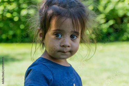 Slika na platnu A young adorable female toddler girl looking past camera with innocent smile and big wide beautiful eyes outside in backyard during summer time
