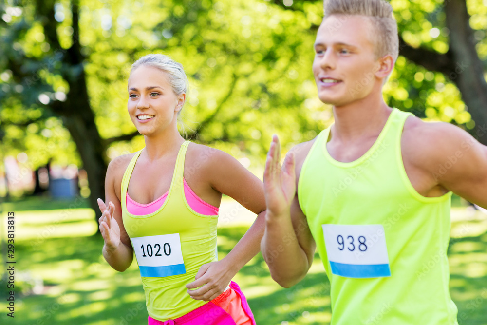 fitness, sport, race and healthy lifestyle concept - happy couple of sportsmen running marathon with badge numbers at summer park fitness, sport, race and healthy lifestyle concept - happy couple of sportsmen running marathon with badge numbers at summer park