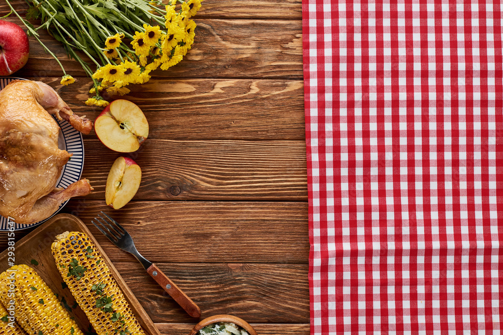 top view of roasted turkey, grilled corn, apples and yellow wildflowers near red checkered napkin on wooden table top view of roasted turkey, grilled corn, apples and yellow wildflowers near red checkered napkin on wooden table