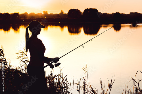Silhouette of a woman at sunset with a fishing rod near the pond.