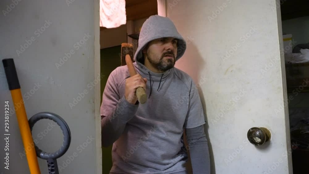 A man in a sweatshirt holds a sledgehammer in his hand. Evil look. Bad intentions. Enters the door. The interior of the garage. Close-up. A man in a sweatshirt holds a sledgehammer in his hand. Evil look. Bad intentions. Enters the door. The interior of the garage. Close-up.