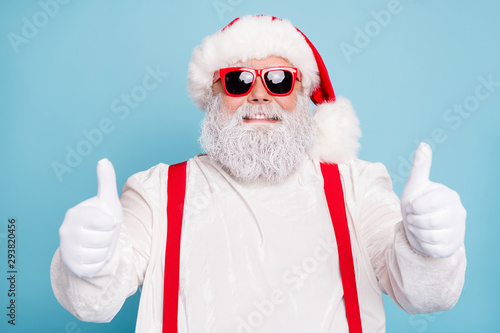 Close up photo of funny funky overweight santa claus in modern spectacles show thumb up sign recommend x-mas season sales wear white overalls red suspenders isolated over blue color background