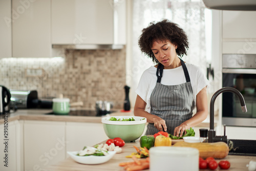 Attractive mixed race woman in apron cutting lettuce for salad. Kitchen interior.