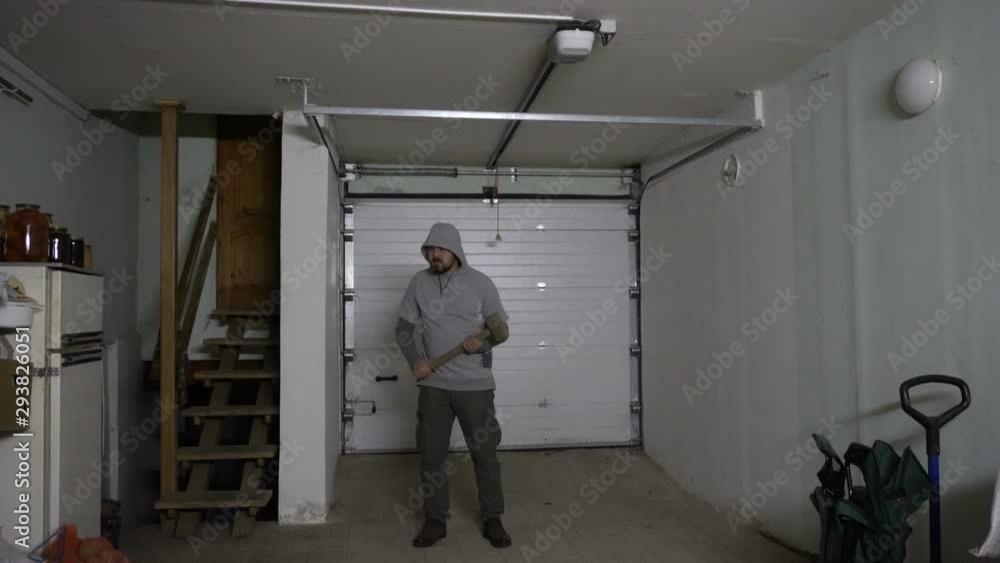 The criminal is moving in the garage. In the hands of an iron sledgehammer. A man is wearing a gray sweatshirt. Against the background of the gate. Professional thief. Maniac in the house. Angry facia The criminal is moving in the garage. In the hands of an iron sledgehammer. A man is wearing a gray sweatshirt. Against the background of the gate. Professional thief. Maniac in the house. Angry facia