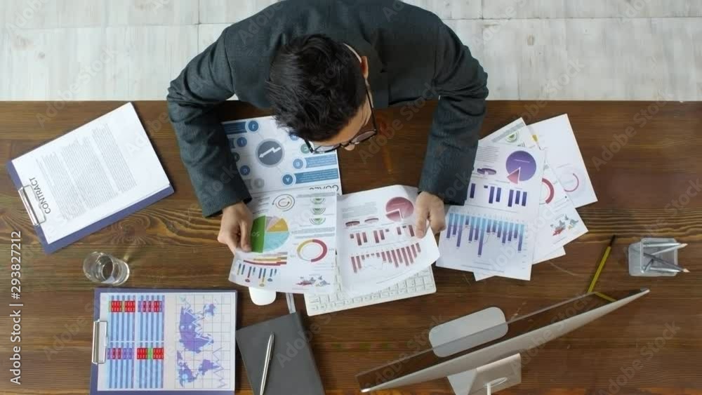 Top view shot of businessman in suit and glasses sitting at table and going through financial documents. He is comparing data and typing on computer Top view shot of businessman in suit and glasses sitting at table and going through financial documents. He is comparing data and typing on computer