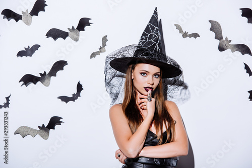 girl in black witch Halloween costume looking at camera near white wall with decorative bats