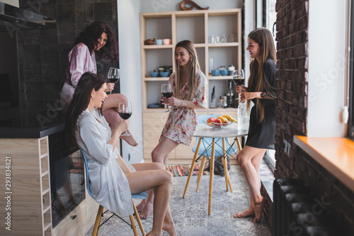Multi ethnic ladies in amazing stylish pajamas enjoy the time together in a modern apartment design holding glasses of wine