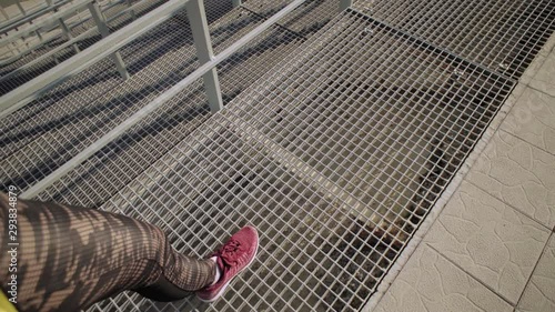 Top view of female legs in red sneakers and dark sportswear, a girl walks on the mesh floor of the stands of an open stadium, tracking shot.