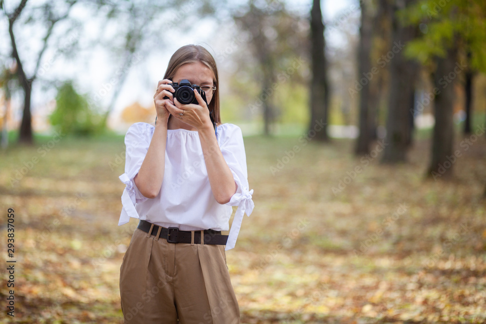 Attractive beautiful young girl holding modern mirror-less camera in autumn park.