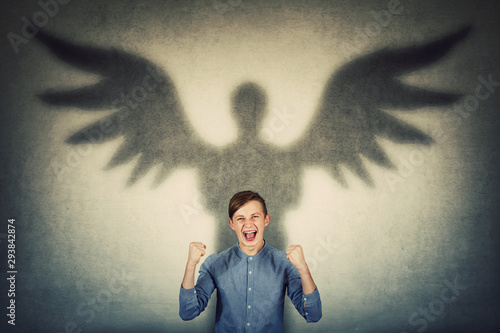 Furious teenage boy holding fists up and screaming showing his powers. Confident adolescent guy casting a superhero shadow with angel wings on a dark room wall. Inner strength and ambition concept. photo