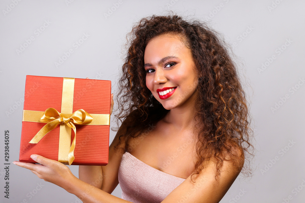 Studio portrait of young woman with dark skin and long curly hair wearing tight sexy dress, holding wrapped present with golden bow. Close up, copy space for text, isolated background. Studio portrait of young woman with dark skin and long curly hair wearing tight sexy dress, holding wrapped present with golden bow. Close up, copy space for text, isolated background.