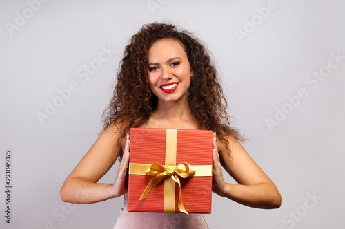 Studio portrait of young woman with dark skin and long curly hair wearing tight sexy dress, holding wrapped present with golden bow. Close up, copy space for text, isolated background. © Evrymmnt
